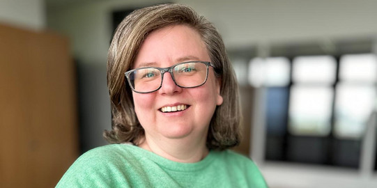 Sandra Szczecina Foto von Sandra Szczecina. Frau mit grünem Pulli, braunen Haaren, einer Brille, lacht in die Kamera.
