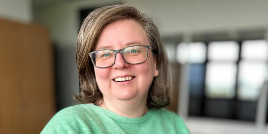 Foto von Sandra Szczecina. Frau mit grünem Pulli, braunen Haaren, einer Brille, lacht in die Kamera.