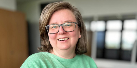 Foto von Sandra Szczecina. Frau mit grünem Pulli, braunen Haaren, einer Brille, lacht in die Kamera.