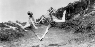 Black and white photo of two women jumping together by hand. One leg is stretched forward and the other leg is bent backward.