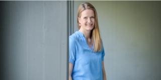 Foto Caterina Schäfer. Frau mit langen blonden Haaren über die linke Schulter gelegt, mit einer hellblauen Bluse an, lächelt Zähne zeigend