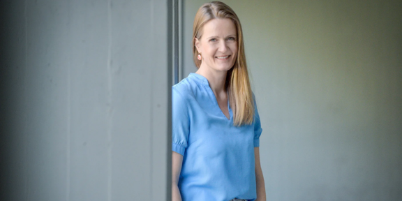 Foto Caterina Schäfer. Frau mit langen blonden Haaren über die linke Schulter gelegt, mit einer hellblauen Bluse an, lächelt Zähne zeigend