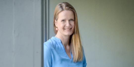 Foto Caterina Schäfer. Frau mit langen blonden Haaren über die linke Schulter gelegt, mit einer hellblauen Bluse an, lächelt Zähne zeigend