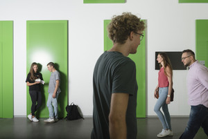 Zwei Studierende lehnen an der Wand, zwei kommen von rechts ins Bild und einer geht nach rechts.